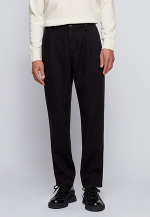 Homme portant un pantalon noir plissé, des chaussures noires à lacets et un pull blanc à manches longues, debout devant un fond gris.