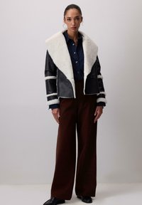 Sieviete ar melnu šerlinga jaku ar baltu vilnas apdari, tumši zilu krekls, plašām brūnām biksēm un melniem apaviem, stāvot pret vienkāršu fona.