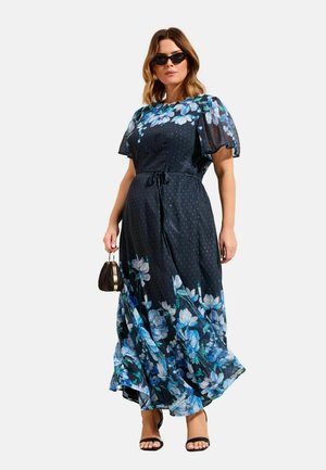 Nő fekete maxiruha visel, kék virágmintával, fodros ujjakkal, fekete napszemüveggel, fekete magassarkú cipővel, és egy kis fekete kézitáskát tart a kezében.