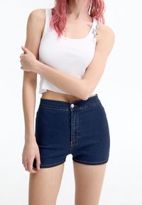 Valkoinen ribatty tank top, jossa on lyhyt malli ja hopeiset yksityiskohdat olkapäässä, yhdistetty tummansinisiin farkkushortseihin, joissa on etupainike.