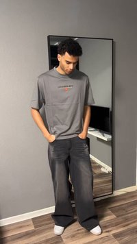 Giovane uomo con capelli ricci in piedi con le mani nelle tasche, indossa una maglietta grigia e jeans neri a gamba larga in una stanza con pavimento in legno e un grande specchio a parete.