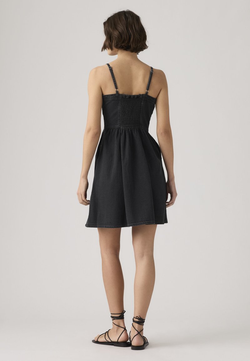 Vrouw met kort haar die een mouwloze zwarte jurk met verstelbare bandjes draagt en zwarte platte sandalen met veters, staand tegen een effen achtergrond.