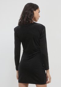 Robe en velours noir à manches longues, avec des motifs de pois dorés, silhouette ajustée et un détail noué à la taille.