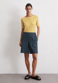 Geel en wit gestreepte short-sleeve top gecombineerd met donkerblauwe shorts met zijzakken. Het model draagt zwarte slidesandalen.