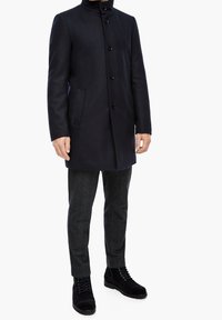 Navy wool blend coat with a high collar, five-button closure, and side pockets, paired with gray textured pants and black lace-up boots.