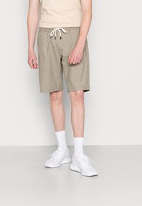Man met beige drawstring shorts, witte crew-sokken en witte sportschoenen, staande tegen een eenvoudige lichte achtergrond.