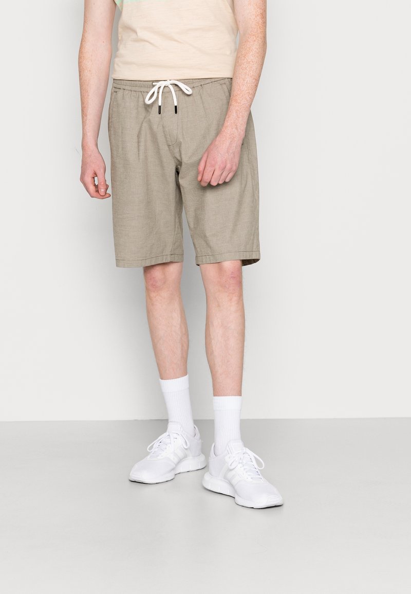 Man met beige drawstring shorts, witte crew-sokken en witte sportschoenen, staande tegen een eenvoudige lichte achtergrond.