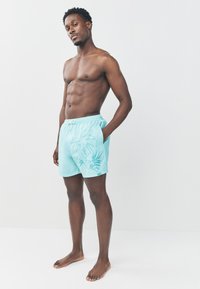 Turquoise zwembroek met een trekkoord in de taille, zijzakken en een palmboomafbeelding. Gemaakt van gladde stof en heeft een ontspannen pasvorm.