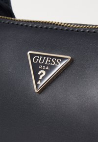 Material preto texturizado com um logo em triângulo realçado a ouro com a inscrição "GUESS U.S.A." apresentando um detalhe de ponto de interrogação e seta.