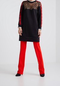 Czarna sukienka z koronkowym górnym wykończeniem, czerwonymi paskami po bokach i długimi rękawami. W zestawie z czerwonymi spodniami i czarnymi butami na szpic.