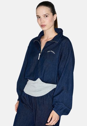 Mujer con chaqueta azul marino con cremallera con texto "Viva Sempre", mangas sueltas y pantalones a juego sobre una camisa gris claro.