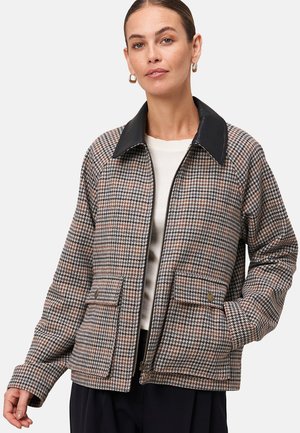 Femme portant une veste zippée en pied-de-poule avec un col noir, des poches à rabat, un haut crème, un pantalon noir et des boucles d'oreilles dorées, posant avec une main dans la poche.