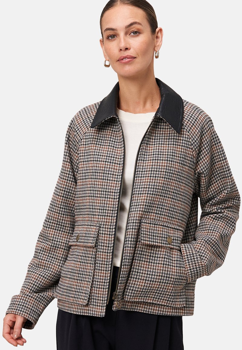 Femme portant une veste zippée en pied-de-poule avec un col noir, des poches à rabat, un haut crème, un pantalon noir et des boucles d'oreilles dorées, posant avec une main dans la poche.