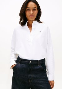 Žena s tmavými vlasy, která má na sobě bílou košili s dlouhým rukávem a malým logem a tmavé vysoký pas džíny, stojí s jednou rukou v kapse.