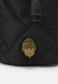 Černá prošívaná látková taška s dekorativním zlatým kovovým zapínáním ve tvaru listu, zdobená malými průhlednými a jantarově zbarvenými kameny.