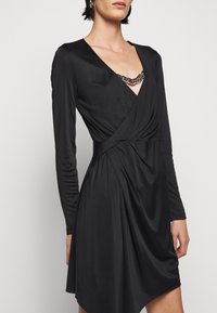 Schwarzes Wickelkleid mit langen Ärmeln, tiefem V-Ausschnitt und einer Kette als Akzent an der Brust. Verfügt über drapierte Stoffe und einen asymmetrischen Saum.