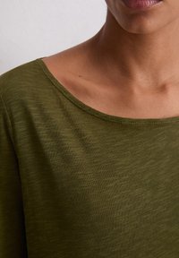 Olive zielony, fakturowany materiał w górnej części z szerokim, zaokrąglonym dekoltem. Materiał wydaje się miękki i lekko elastyczny, z subtelnymi różnicami w odcieniu.