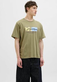 Olivengrøn bomulds t-shirt med korte ærmer, der har et flerfarvet logodesign på forsiden, parret med mørk denim bukser.