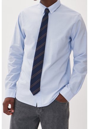 Homme portant une chemise bleue claire boutonnée avec une cravate rayée bleu foncé et marron et un jean gris, une main dans la poche, debout.