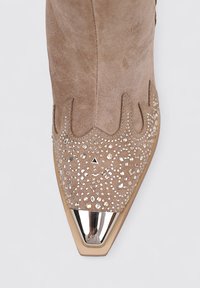 Botín de ante en marrón claro con strass decorativos y puntera metálica puntiaguda. Presenta bordes ondulados y una superficie texturizada.