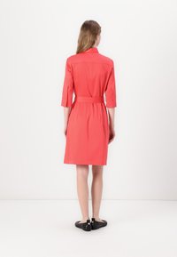 Coral dress with three-quarter sleeves, a collar, and a belted waist. Soft fabric with a smooth texture. Black shoes with a decorative element.