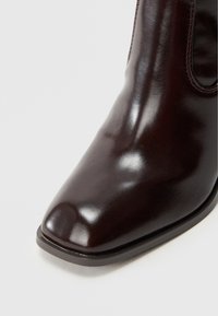 Bordowy skórzany botek za kostkę z błyszczącym wykończeniem, szpiczastym noskiem, elastycznymi panelami po bokach oraz płaską, teksturowaną podeszwą.