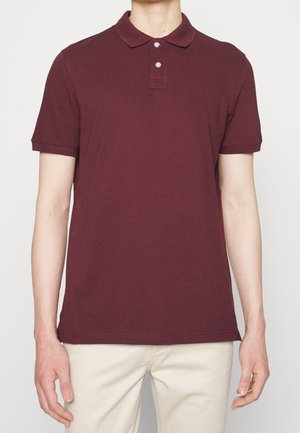 Mand iført en kortærmet mørkerød polo med to knapper, kombineret med lyse beige bukser, vist fra nakke til hofter.