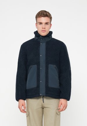 Nuori mies, jolla on yllään tummansininen teksturoitu fleece-takki korkealla kauluksella ja vaaleat cargo-housut, seisoo valkoista taustaa vasten.