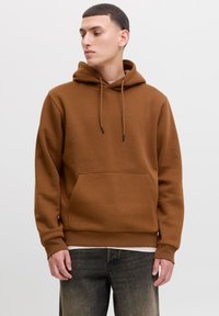 Ung mand iført en brun hættetrøje og mørke jeans, der kigger til venstre mod en ensfarvet lys baggrund.