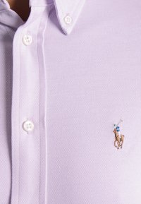 Camisa polo de color púrpura claro, con tejido texturizado, botones blancos y un pequeño logo bordado de un caballo y un jinete en marrón y azul.