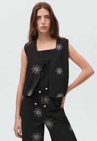 Musta hihaton toppi V-pääntiellä ja etusolmulla, jossa on hopeista tähtipurske-ompelua. Pehmeä kangas, jossa on rento istuvuus.