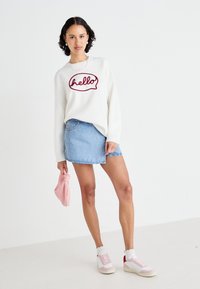 Hvit hettegenser med "hello" i burgunder, kombinert med en lys blå denimskjørt. Modellen holder en lys rosa veske. Har på seg casual fottøy.