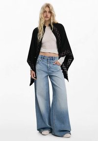 Sciarpa nera con ricamo a zigzag rosa, indossata sopra un top bianco corto e jeans a gamba larga azzurri. Sono evidenziate texture e forme distintive.