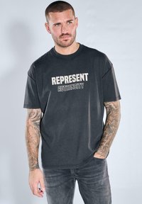 Man with tattoos wearing a black T-shirt with "Represent Authencity" text, standing with one hand in pocket against a plain light background.