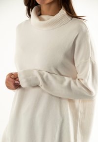 Suéter de cuello alto color crema con un cuello acanalado, textura suave y corte oversize, que cuenta con mangas largas y puños ajustados.