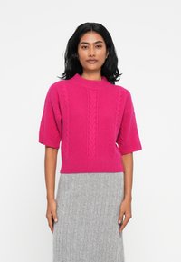 Pull en tricot rose vif à manches courtes et col montant. Présente des motifs en torsades et un ourlet côtelé. Associé à une jupe grise.