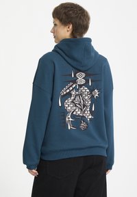 Szmaragdowy hoodie z dużą czarno-białą grafiką na plecach, przedstawiającą abstrakcyjne wzory, oczy i motywy łez. Miękki materiał, luźny fason.
