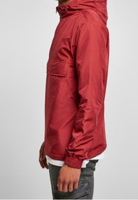 Mężczyzna w czerwonej kurtce przeciwwiatrowej z kapturem, z elastycznymi mankietami i kieszenią z przodu, w połączeniu z białą koszulą i ciemnoszarymi spodniami.