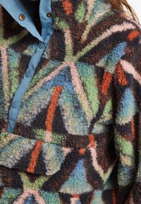 Fleece-paita monivärisessä geometrisessä kuosissa, pehmeällä tekstuurilla, sinisessä napitettavassa kauluksessa ja edessä taskussa logotunnuksella.