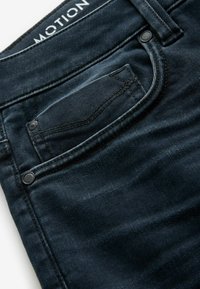 Jean en denim bleu foncé avec poche avant incluant une petite poche à monnaie, rivets métalliques et étiquette "MOTION" visible à l’intérieur de la ceinture.