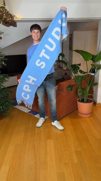 Lichtblauwe sjaal met witte tekst "CPH STUDIO," met franjes aan de randen, vastgehouden door een persoon in casual kleding. Binnenomgeving met planten en meubels.