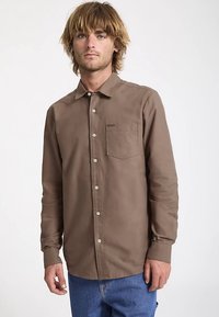 Chemise à manches longues boutonnée en coton brun, dotée d'une poche poitrine et de boutons de couleur claire, portée sur un jean bleu.