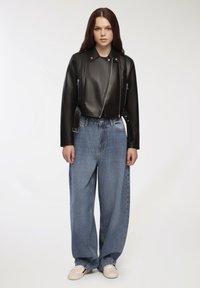 Giacca cropped nera in pelle con zip metalliche, abbinata a jeans di denim blu a vita alta e larghi. Modello in piedi su uno sfondo neutro.