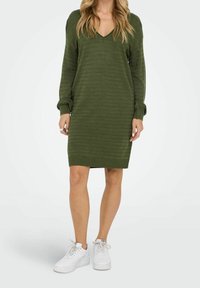 Robe pull verte tricotée avec encolure en V, manches longues, poignets et ourlet côtelés. Associée à des baskets blanches pour un look décontracté.