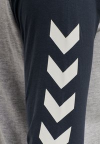 Nærbillede af en ærme med fire hvide chevronpile, der peger nedad, på et mørkeblåt stof ved siden af en grå stofbaggrund.