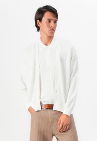 Man wearing white buttoned cardigan over white polo shirt, beige ribbed trousers, and brown leather belt, posing against white background.