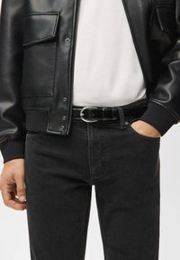 Schwarze Lederjacke mit Druckknöpfen, schwarze gerippte Bündchen; darunter weißes T-Shirt, kombiniert mit schwarzer Denim-Jeans und einem Ledergürtel mit metallischer Schnalle.