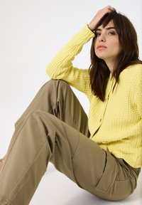 Gehaakte gele cardigan met een textuurpatroon, knopen aan de voorkant, gecombineerd met lichtbruine, losse cargo-achtige broeken.
