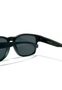 Occhiali da sole con montatura nera e lenti scure, caratterizzati da una finitura opaca e un design rettangolare elegante. "Fatto in Spagna" stampato sul braccio.