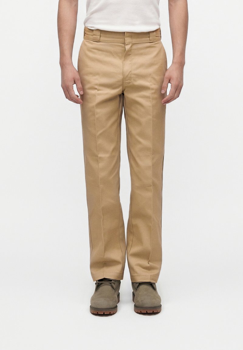 Man draagt beige rechte broek, bruine veterlaarzen en een wit mouwloos top tegen een effen witte achtergrond.